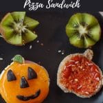 Pinterest pin with white text on black background and photo of Halloween Breakfast Pizza Sandwich cut into a jack o'lantern on a black plate with kiwi flowers