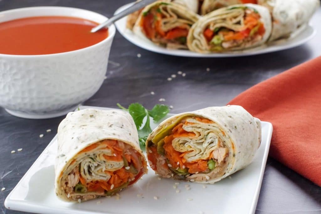 healthy turkey wrap, sliced in half, on white plate, with bowl of tomato soup and platter of turkey wraps in the background