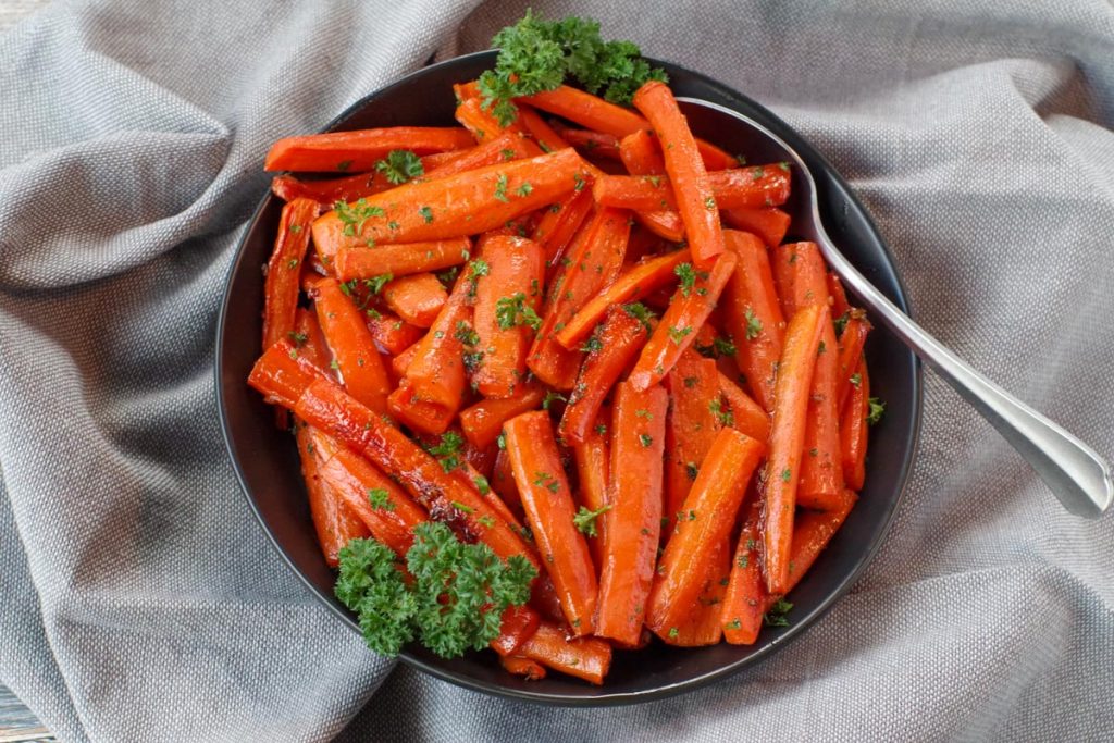 brown sugar glazed carrots in a black bowl, with a spoon, on grey linen