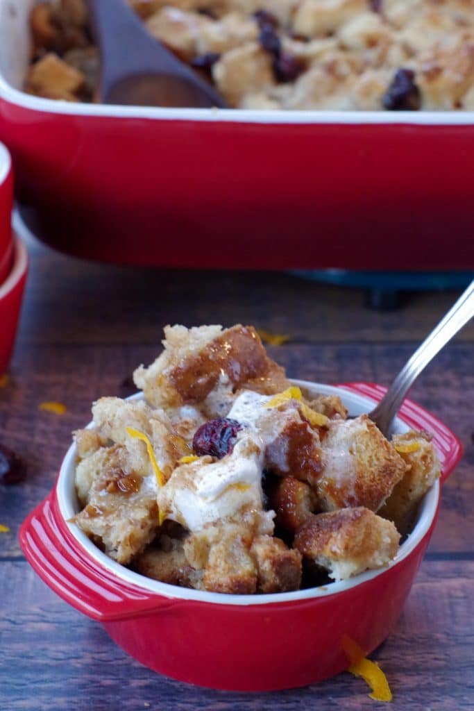 cranberry toffee bread pudding in a red bowl with a spoon and red pan of bread pudding in the background