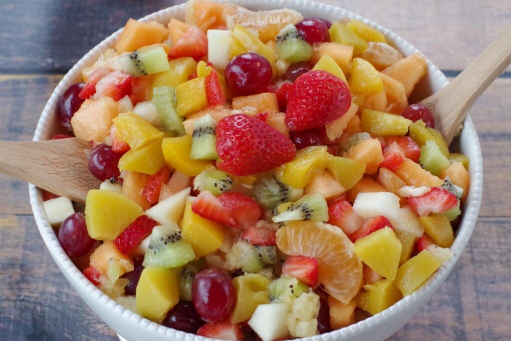 classic fruit salad in a white bowl with wooden serving spoons on each side (on faux wooden surface)