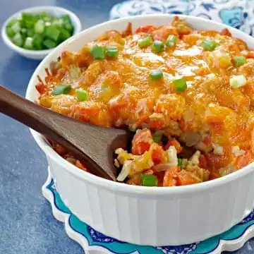 carrot casserole in white casserole dish with a brown wooden spoon scooping casserole (on blue background) with dish of green onions and oven mitts in the background
