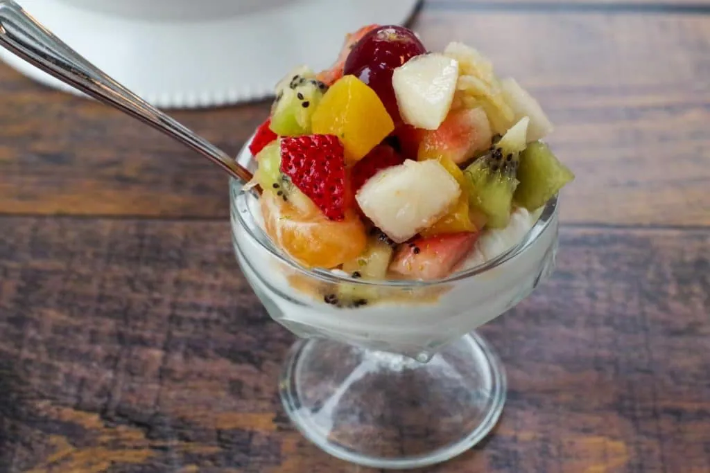 fruit salad over Greek yogurt in a glass dessert dish, with a spoon (on faux wooden surface)