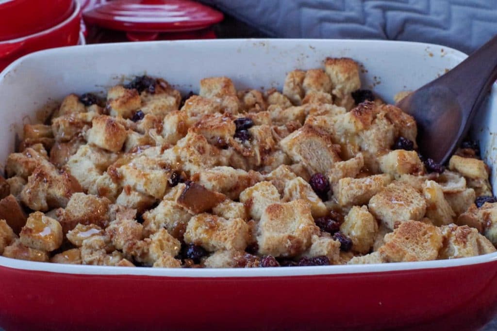 bread pudding with condensed milk in a red casserole dish the a wooden spoon in it and red dishes and grey oven mitts in background