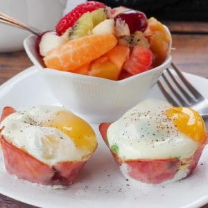 healthy eggs benedict on a white plate with a fork and a bowl of fruit