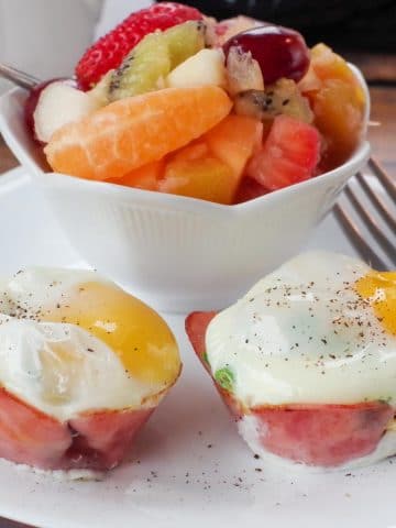healthy eggs benedict on a white plate with a fork and a bowl of fruit