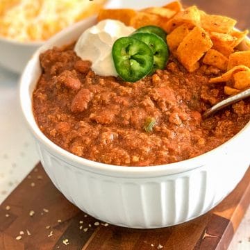 chili in a white casserole bowl, topped with jalapenos, sour cream and chips, on a brown cutting board