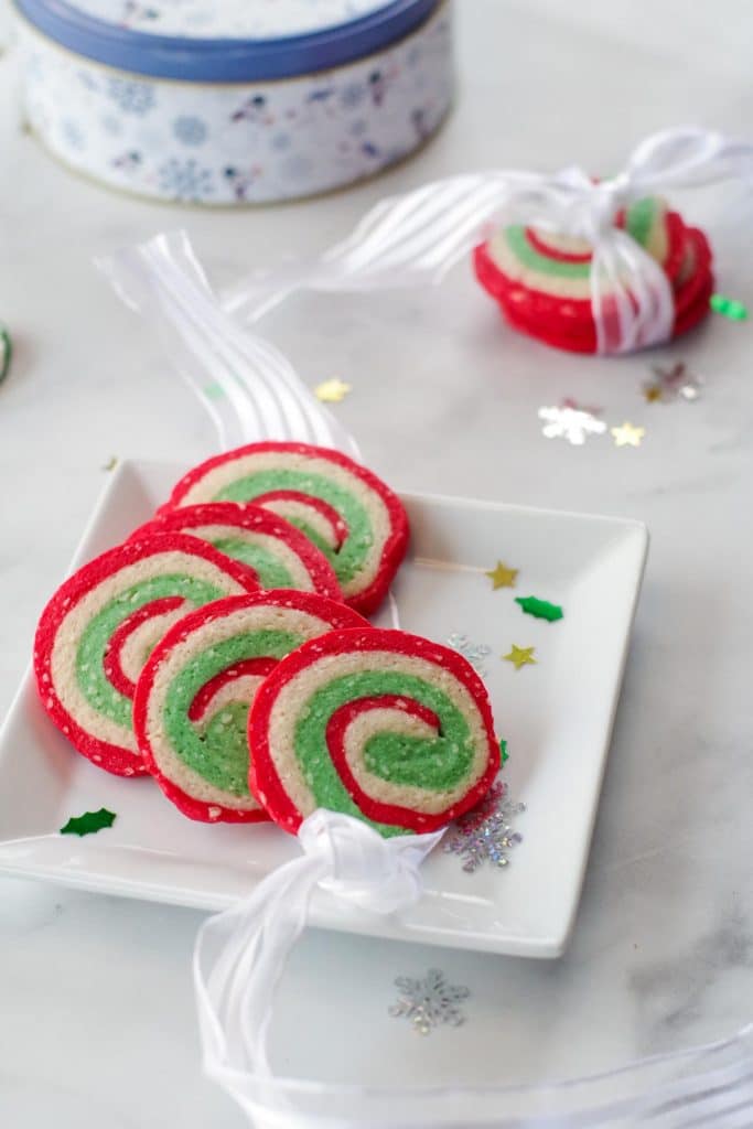 4 Christmas pinwheel cookies on a white plate with 3 cookies in a ribbon in the background, as well as a cookie tin