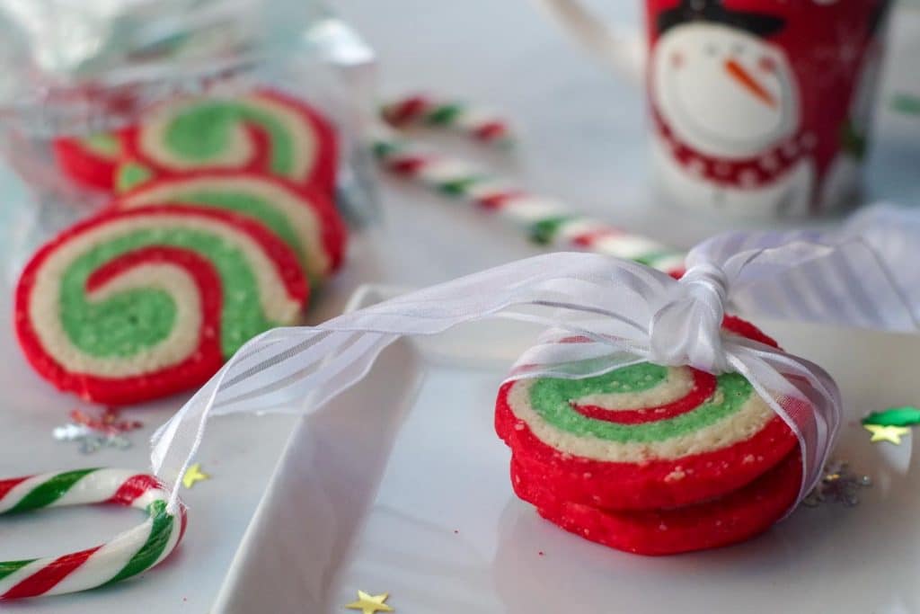 3 Christmas swirl cookies on a white plate, tied with a white bow, with more cookies, a candy cane and a mug in the background