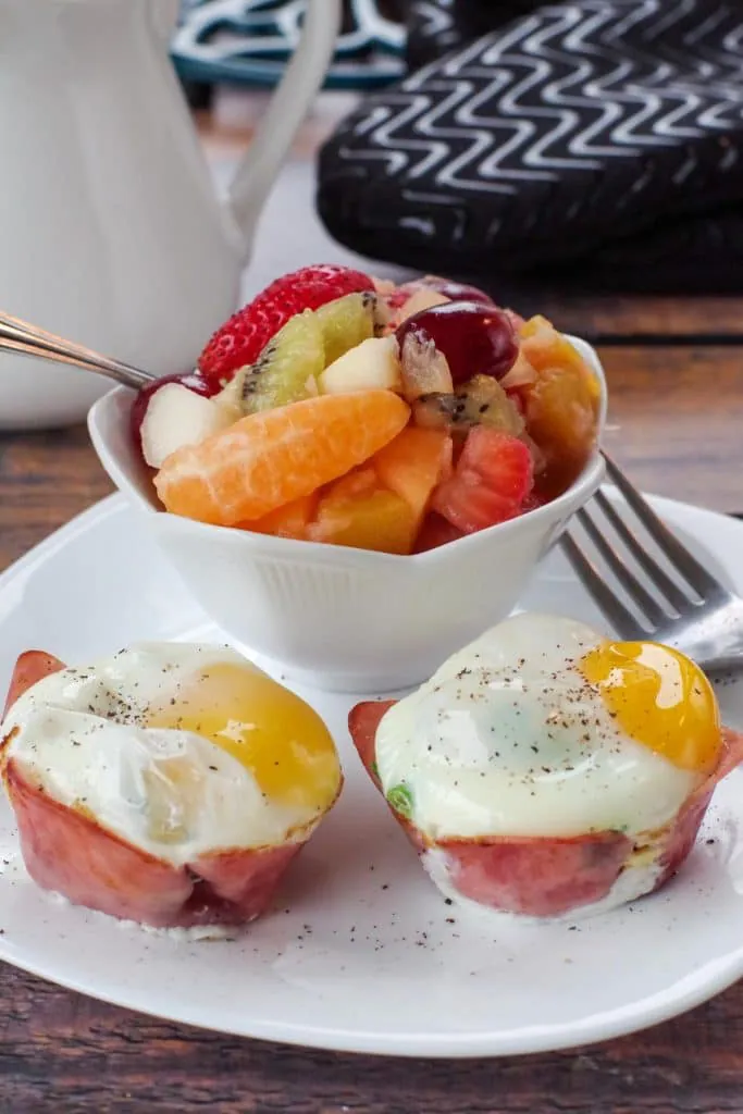 2 healthy eggs benedict on a white plate with a fork and a bowl of fruit