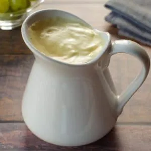Healthy Hollandaise sauce in a large white creamer, with a bowl of green grapes in background and grey linen