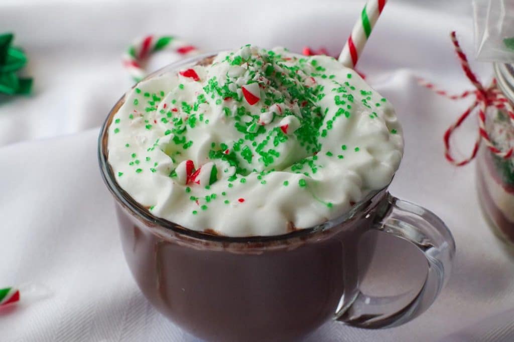 candy cane hot chocolate in a clear mug with whipped cream, green sprinkles and crushed candy canes