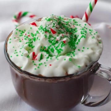 candy cane hot chocolate in a clear mug with whipped cream, green sprinkles and crushed candy canes