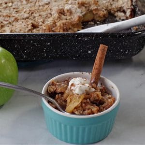 apple crisp in a blue container with a pan of apple crisp in the background