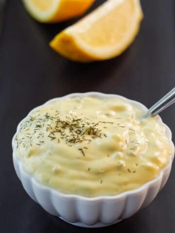 tartar sauce in a small white dish with a spoon sticking out of it, sitting on a black tray with sliced lemon in the background