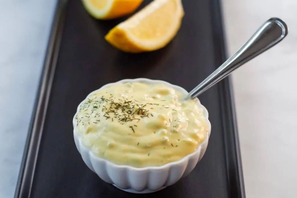 tartar sauce in a small white dish with a spoon sticking out of it, sitting on a black tray with sliced lemon in the background