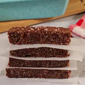 4 No bake energy bars stacked with parchment paper between them and blue loaf pan on cutting board in background