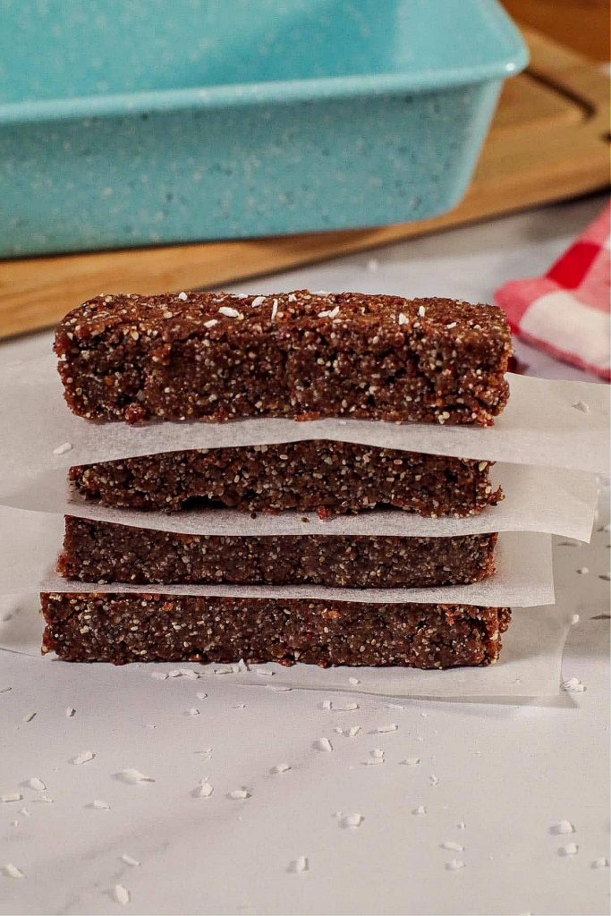 4 No bake energy bars stacked with parchment paper between them and blue loaf pan on cutting board in background