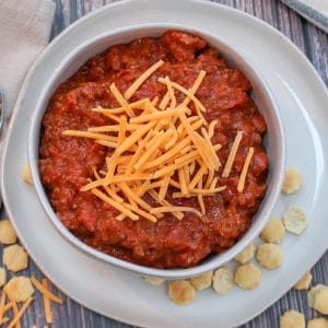 tailgate chili in a white bowl on a platter, with cheese on top and little crackers scattered around