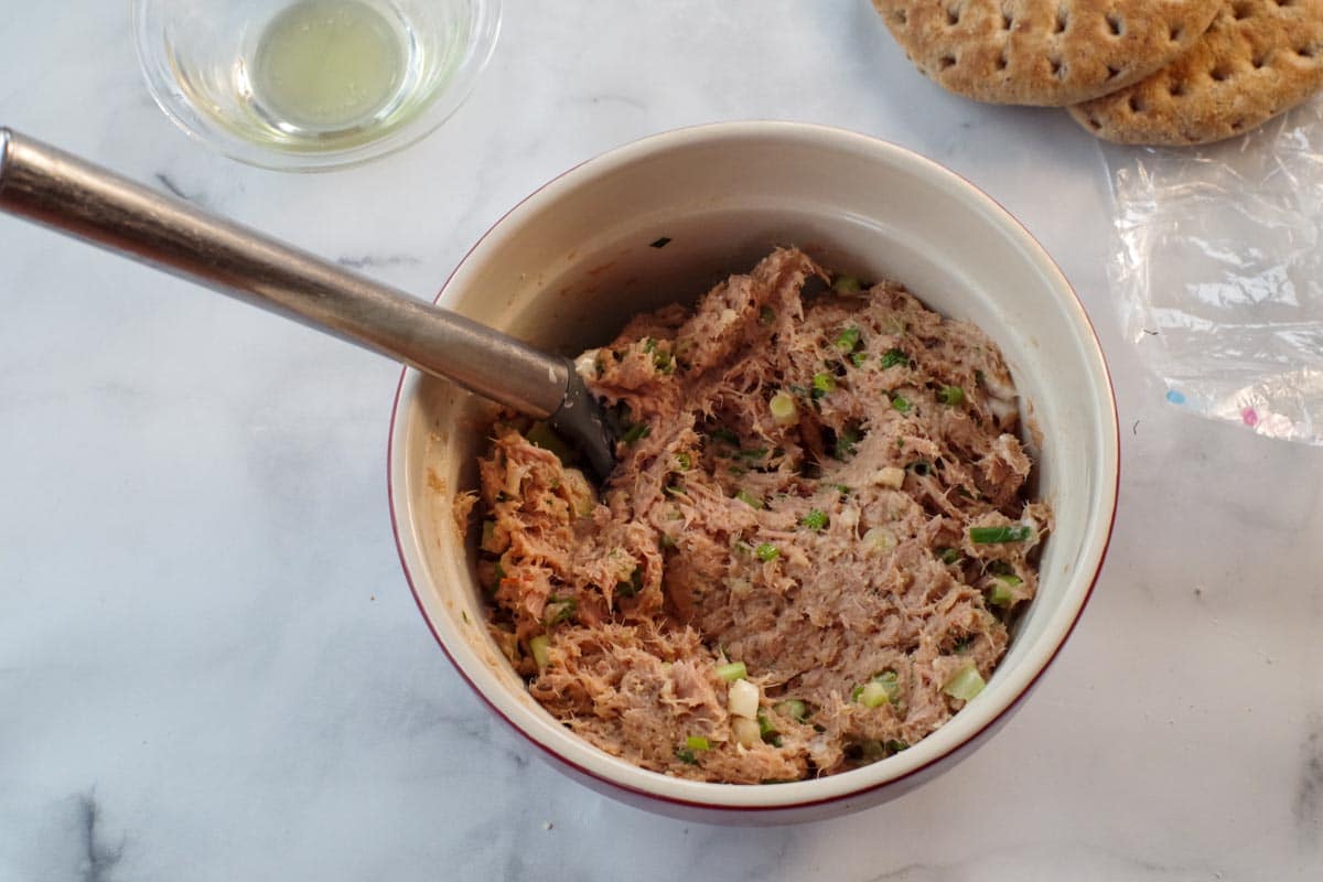 tuna burger ingredients mixed together in a bowl with a spoon and olive oil in the background