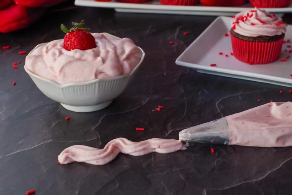 strawberry whipped cream frosting in a bowl with a strawberry, a pastry bag with a line of frosting coming out and a frosted cupcake in the background