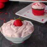 strawberry frosting in white bowl with cupcake on a plate in the background
