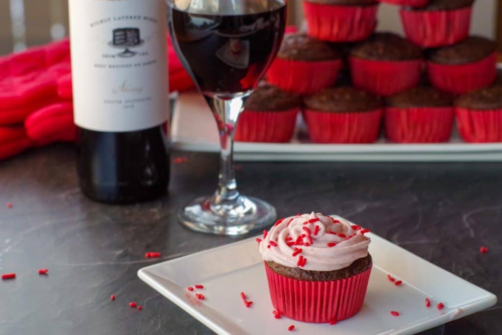red wine cupcake on a white plate with bottle of wine, a glass of wine and atray of cupcakes stacked in the background