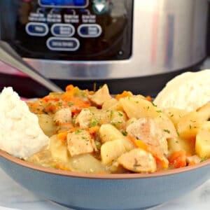 crockpot chicken stew in a blue bowl with biscuits on the side and a slow cooker in the background