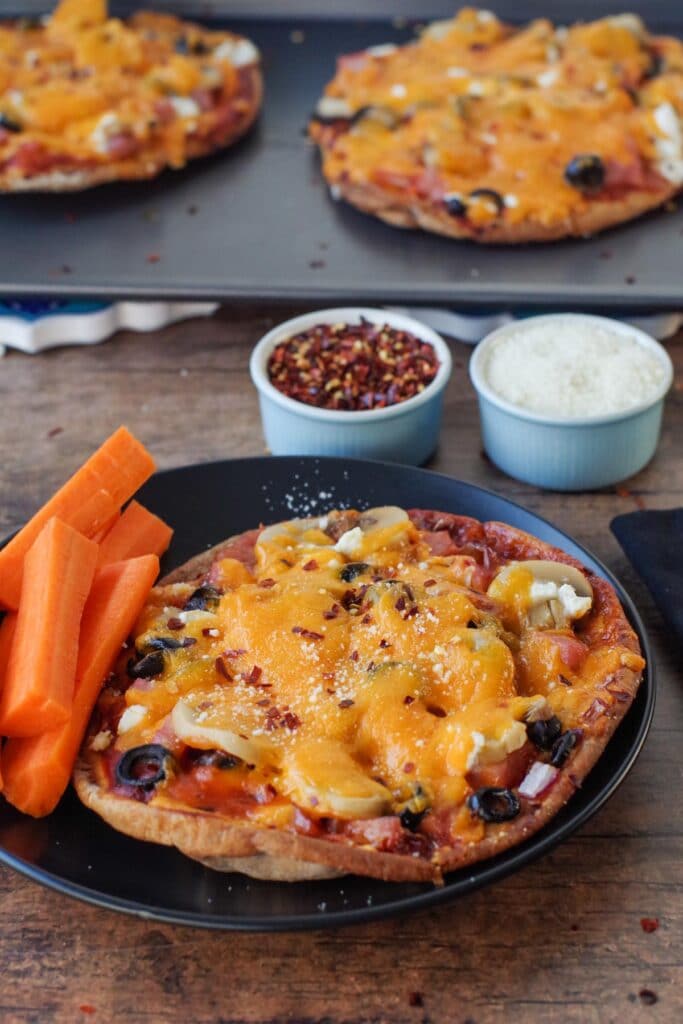 pita pizza on black plate with carrot sticks and more pizzas on baking sheet in background