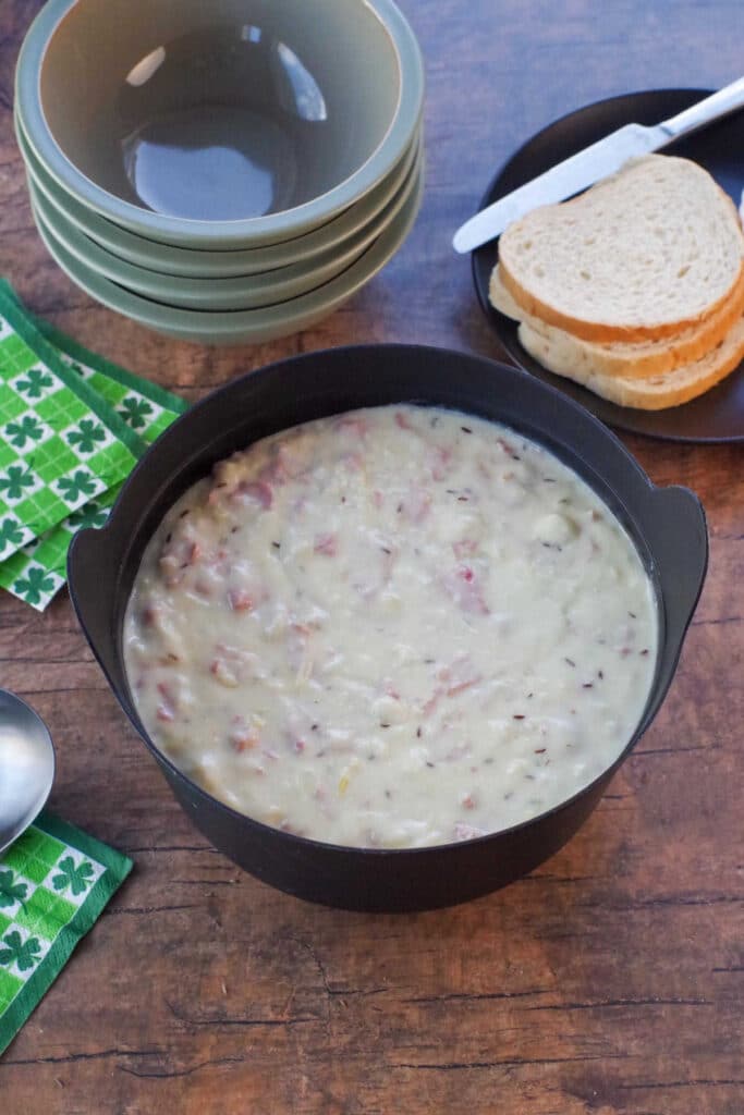 Reuben chowder in a black pot with ladle and clover leaf green and white napkins, sage green bowls and rye bread in the background