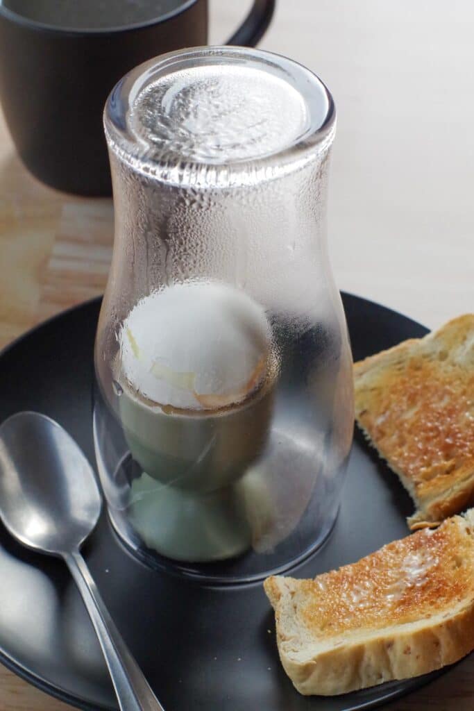 soft-boiled egg under a glass with toast on the side and spoon on other side