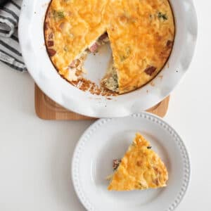 a piece of crustless ham and cheese quiche on a white plate and a whole quiche (with a piece missing) in the background