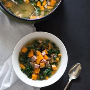 Sweet potato ham and kale soup in a white bowl with a spoon and a pot of soup behind