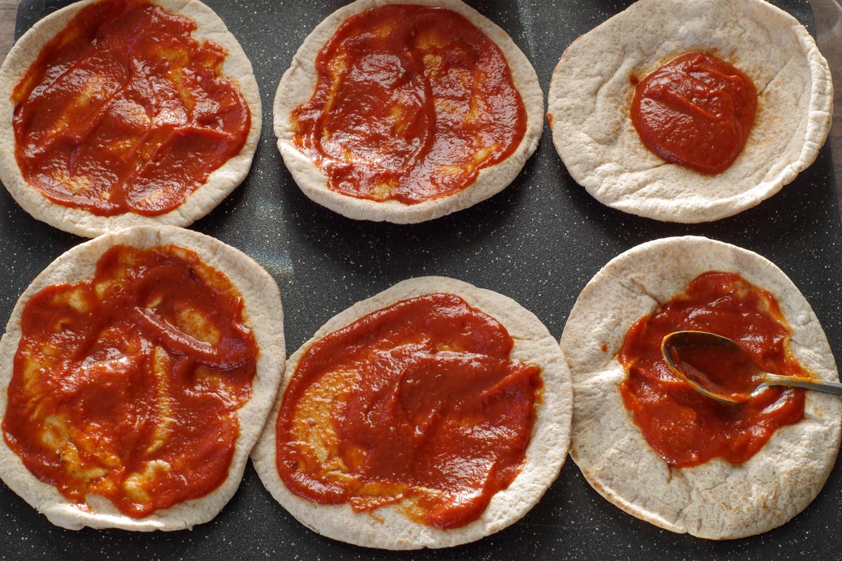 6 pitas on cutting board with pizza sauce being spread on them