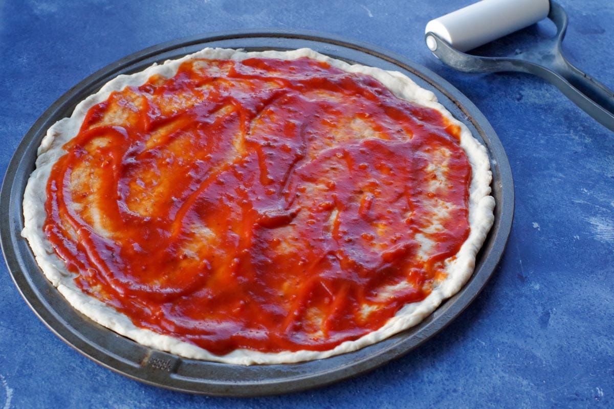 pizza dough with sauce on pan, on blue counter top surface with rolling pin in background