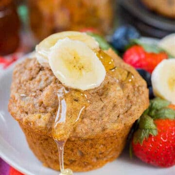 banana bran muffin with banana slices on top and syrup dripping off, on white plate with strawberries on the side