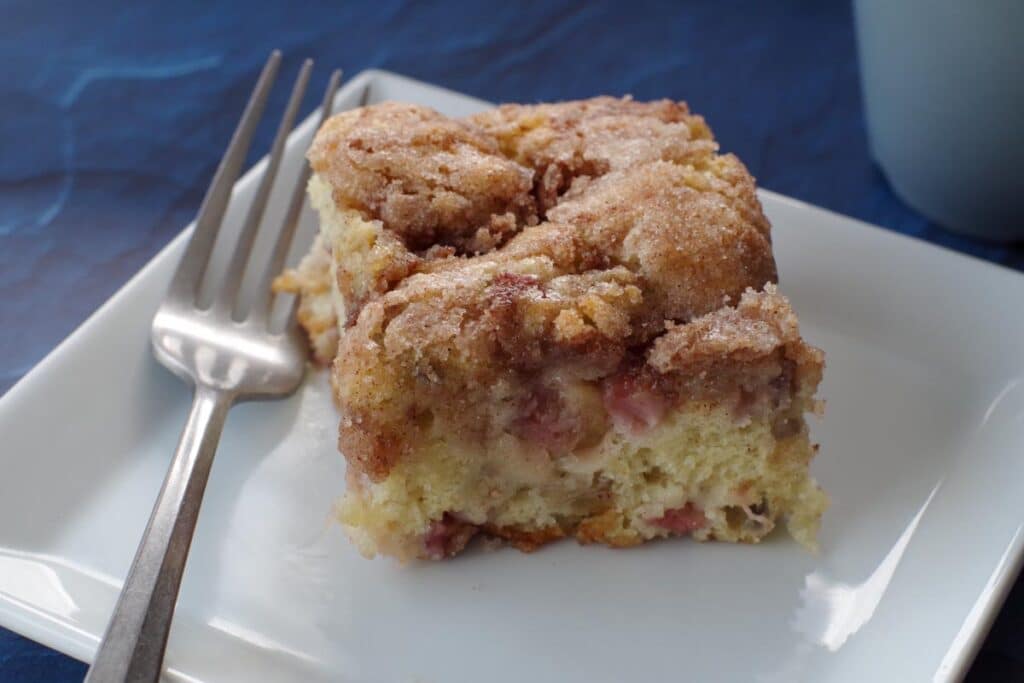 piece of rhubarb coffee cake on a white plate with a fork on the side, on a blue surface