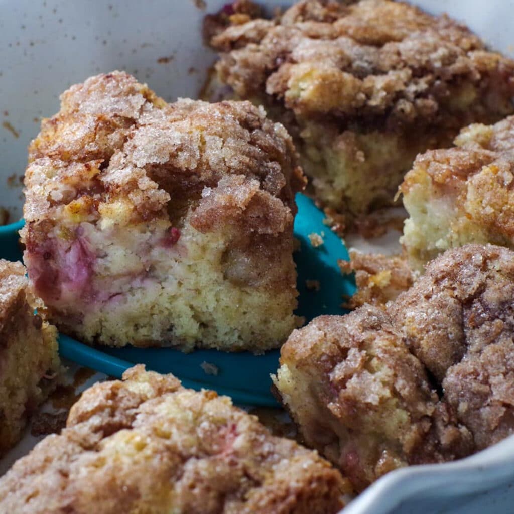 rhubarb coffee cake cup up, in off-white baking dish, with piece being lifted out with blue spatula