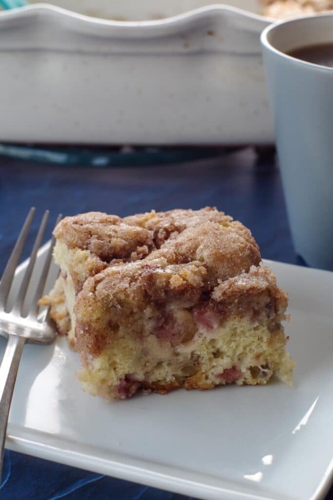 piece of rhubarb coffee cake on a white plate with a fork on the side, on a blue surface