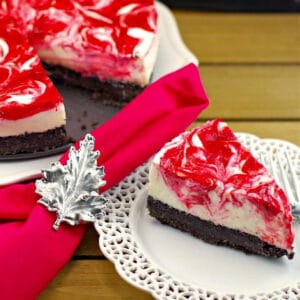 a piece of strawberry rhubarb cheesecake on a white plate with a red napkin with Canadian flag napkin ring and remainder of cake in the background