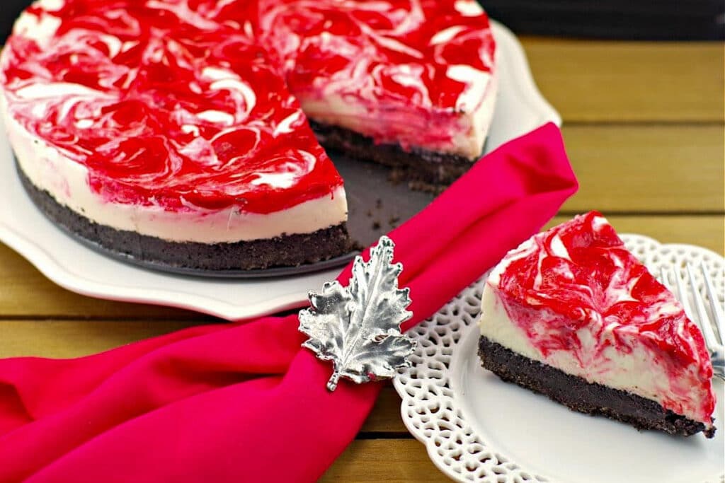 a piece of strawberry rhubarb cheesecake on a white plate with a red napkin with Canadian flag napkin ring and remainder of cake in the background