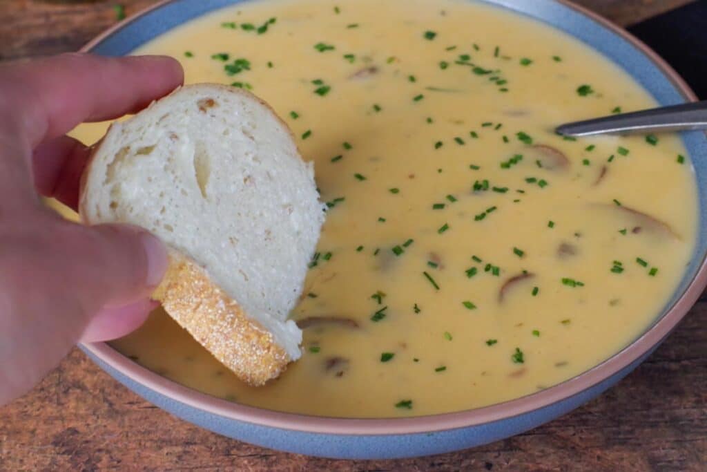 piece of bread being dipped into a bowl of beer cheese soup with a spoon in it