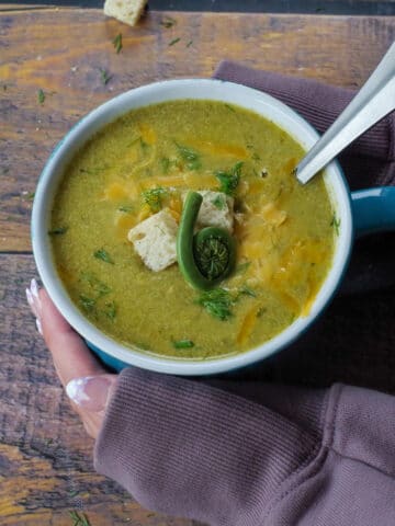 fiddlehead soup in a mug with a spoon in it and hands cradling it