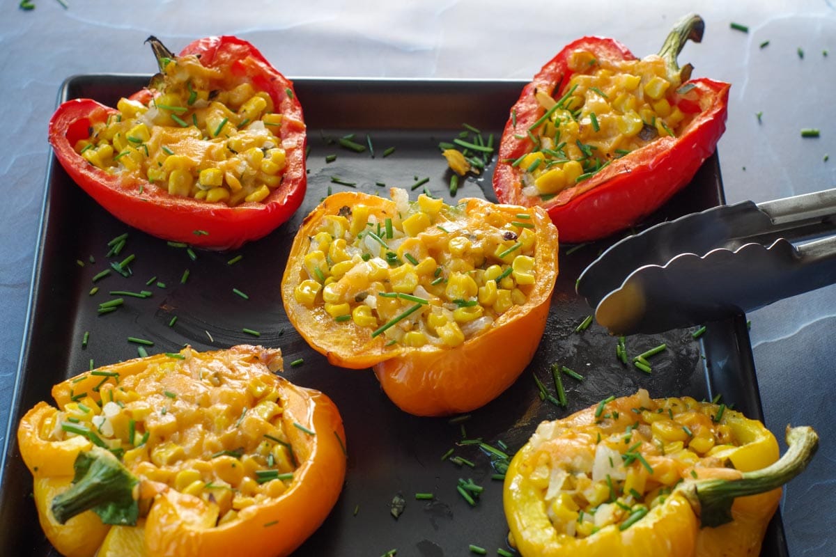 5 Stuffed grilled peppers on a black platter with tongs leaning on platter