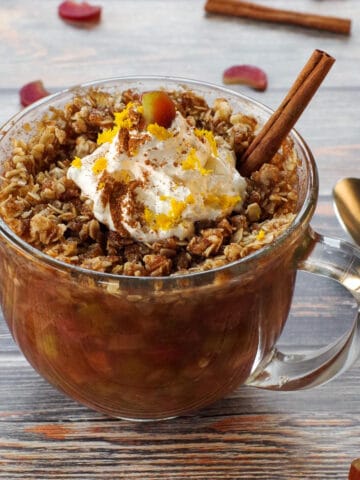 rhubarb crisp in a mug, topped with whipped cream with a stick of cinnamon, a spoon on the side and chopped rhubarb and cinnamon sticks strewn around