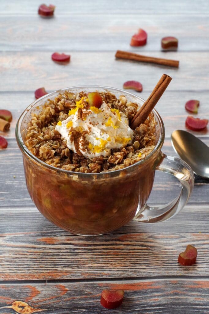 rhubarb crisp in a mug, topped with whipped cream with a stick of cinnamon, a spoon on the side and chopped rhubarb and cinnamon sticks strewn around