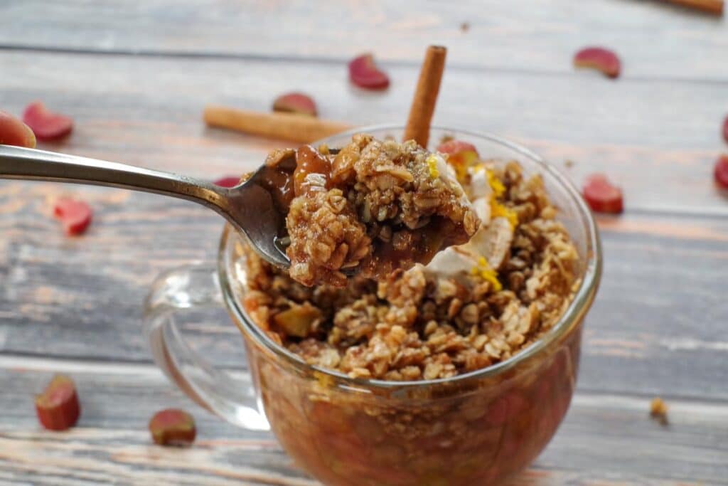 rhubarb crisp in a mug, topped with whipped cream with a stick of cinnamon, a spoon being held up with rhubarb crisp on it, and chopped rhubarb and cinnamon sticks strewn around
