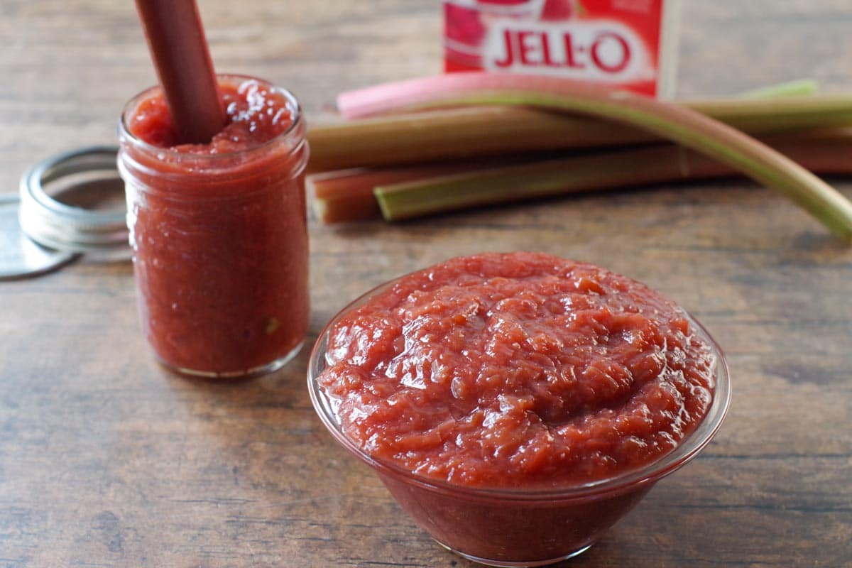 stewed rhubarb in glass bowl with jar of stewed rhubarb and fresh rhubarb and a box of strawberry jello in the background.