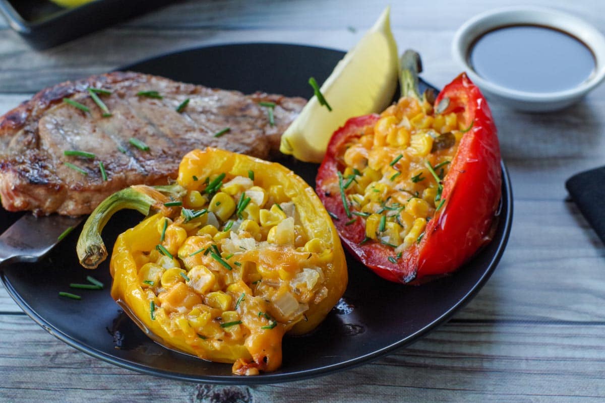 2 stuffed grilled peppers on a black plate with a piece of grilled pork and lemon wedge