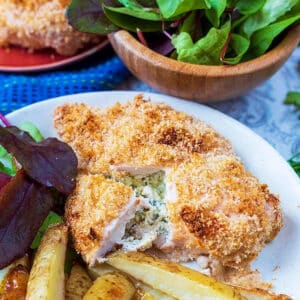 chicken kiev on a plate with potato wedges and a bowl of greens in the background